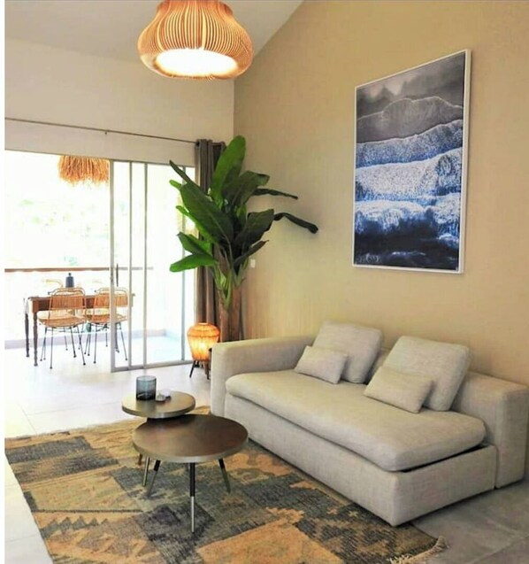 A modern living room with a sofa, a coffee table, and a large plant. There is a painting on the wall and a dining area in the background.