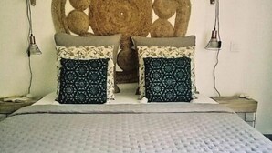 Un lit avec une couette grise, deux coussins à motifs, une tête de lit en osier et un livre intitulé « MAISONS CRÉOLES » posé sur un tapis en fibres tressées.