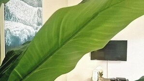 Un salon avec une grande plante verte au premier plan, un canapé blanc, un téléviseur à écran plat accroché au mur et une petite table sur laquelle se trouvent une lampe et un ananas.