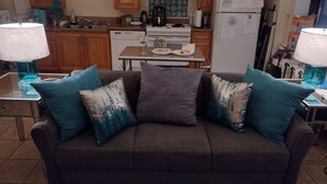 Uma sala de estar com um sofá marrom, almofadas verde-água, uma mesa de centro e uma cozinha ao fundo.