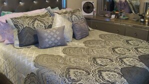 Quarto com cabeceira estofada, roupa de cama estampada, um ventilador de cabeceira e um espelho refletindo o aposento.