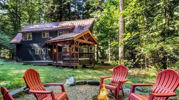 A cabin with a deck, surrounded by a fire pit and red Adirondack chairs. - Wild Goose Lodge | Hot Tub + Bunk Room (Glacier)