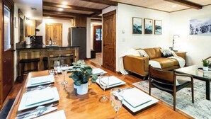 A dining area with a wooden table set for a meal, a sofa, and a kitchen in the background. - Wild Goose Lodge | Hot Tub + Bunk Room (Glacier)