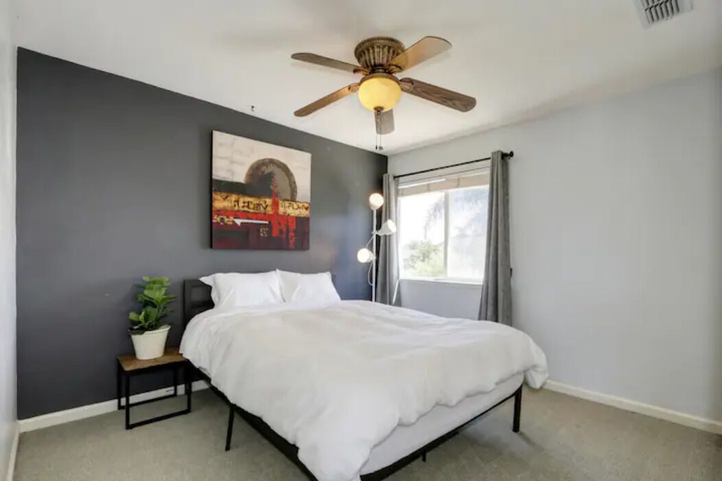 Un dormitorio con cama, ventilador de techo, ventana con cortinas, mesita de noche con planta y una obra de arte colgada en la pared.