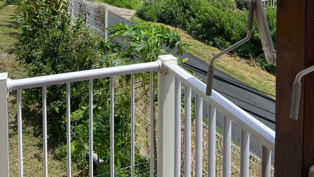 Un balcone con vista su una strada e del verde, con ringhiera bianca e recinzione in metallo.