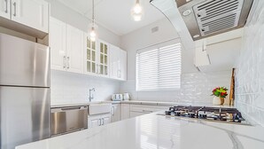 A modern kitchen with white cabinetry, stainless steel appliances, a marble island, and a window with blinds. - The Cottage - Lithgow (Lithgow)
