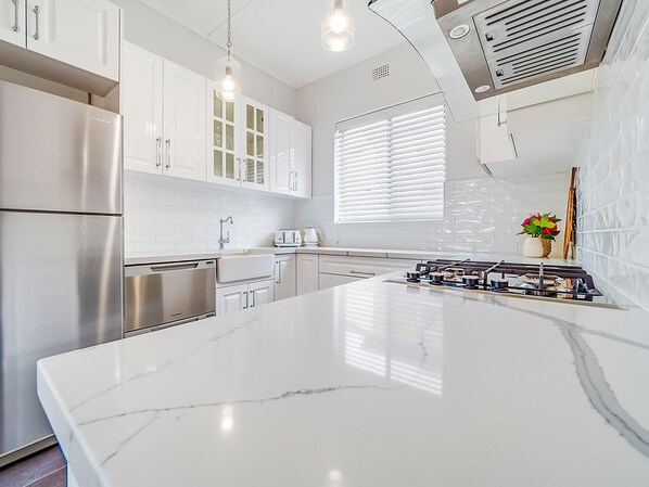 A modern kitchen with white cabinetry, stainless steel appliances, a marble island, and a window with blinds. - The Cottage - Lithgow (Lithgow)