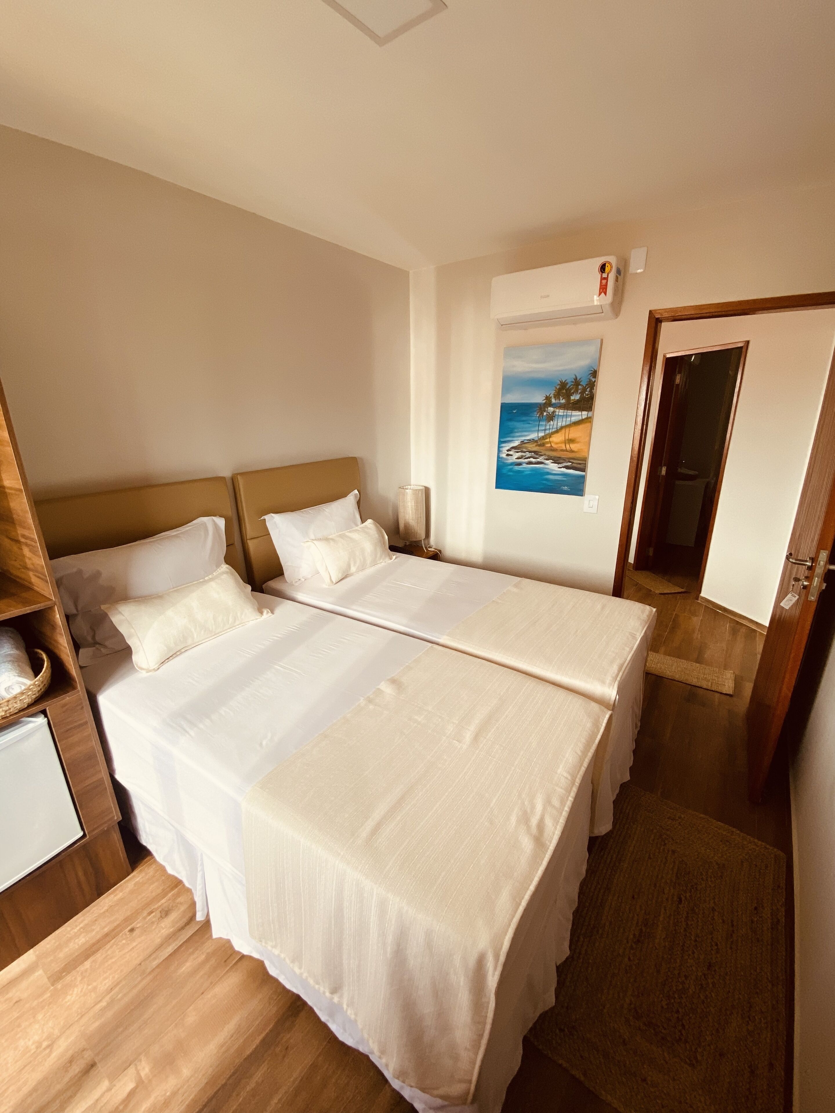 A hotel room with two beds, a painting of a beach scene, a wooden floor, and a door to the right.