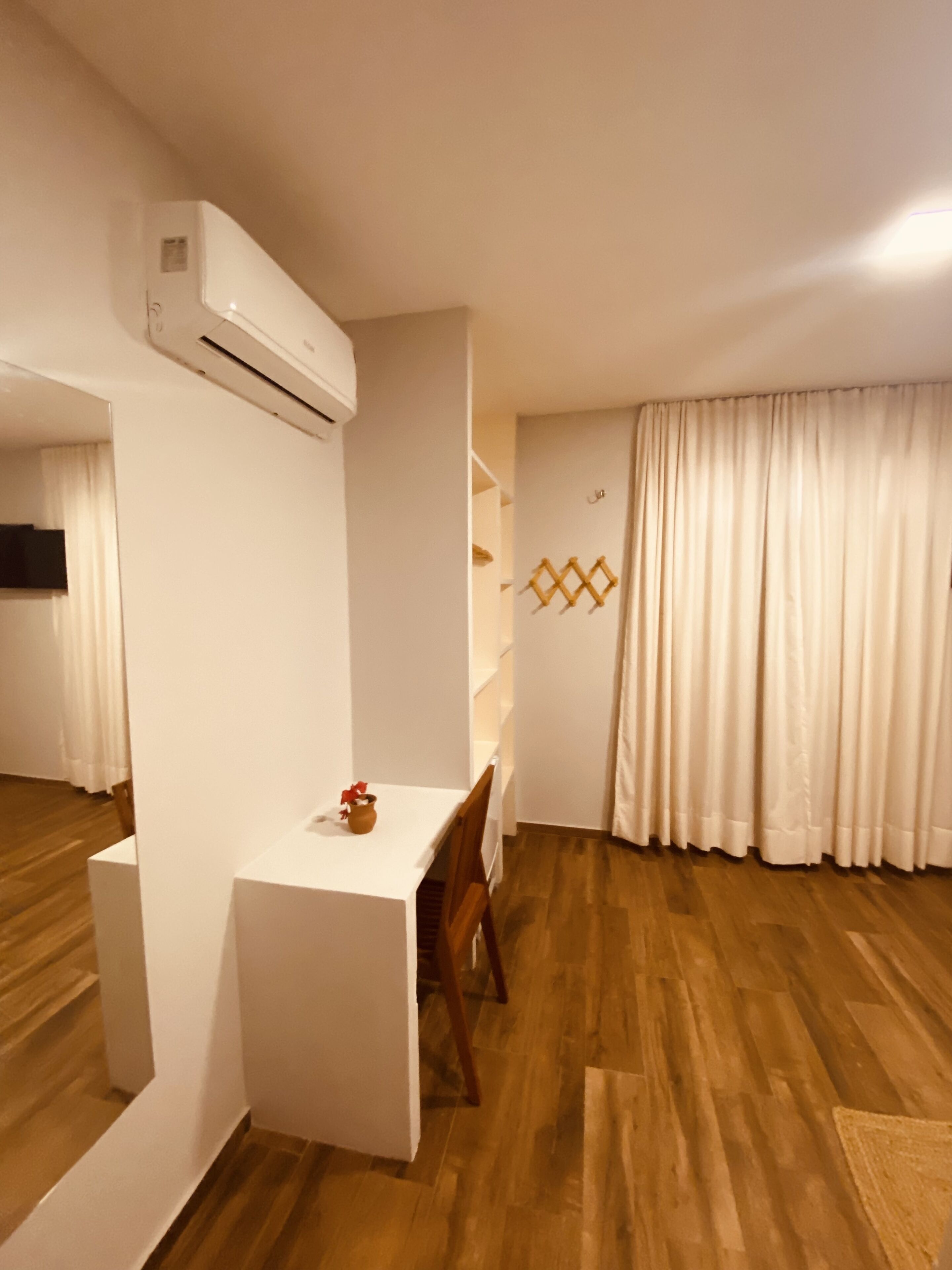 A room with a wooden floor, a white table with a chair, a white air conditioning unit, and a window with white curtains.