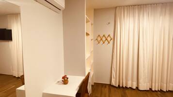 A room with a wooden floor, a white table with a chair, a white air conditioning unit, and a window with white curtains.