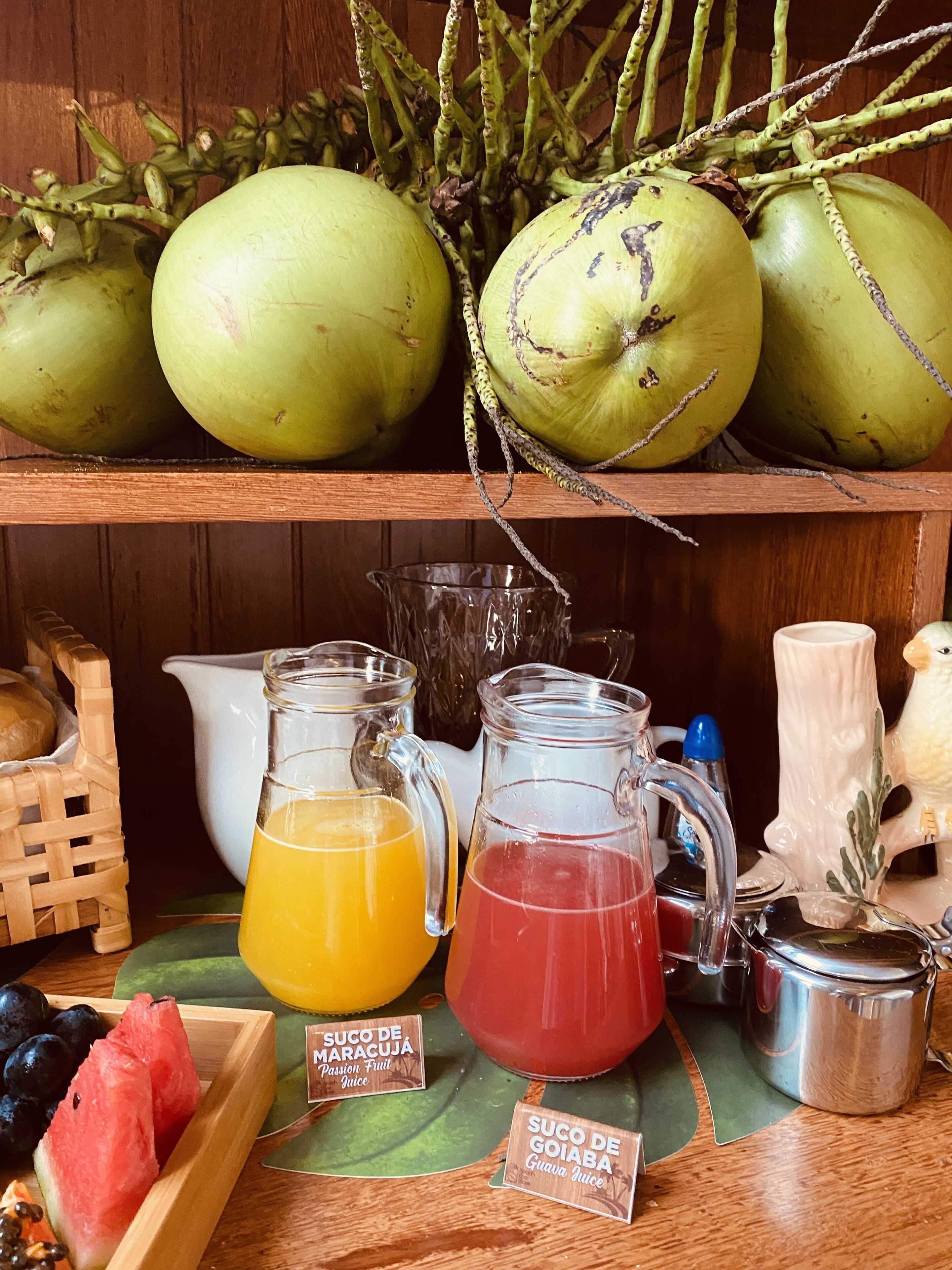 A wooden shelf with coconuts, two glass pitchers of juice, a white pitcher, and a wooden bowl with watermelon slices.