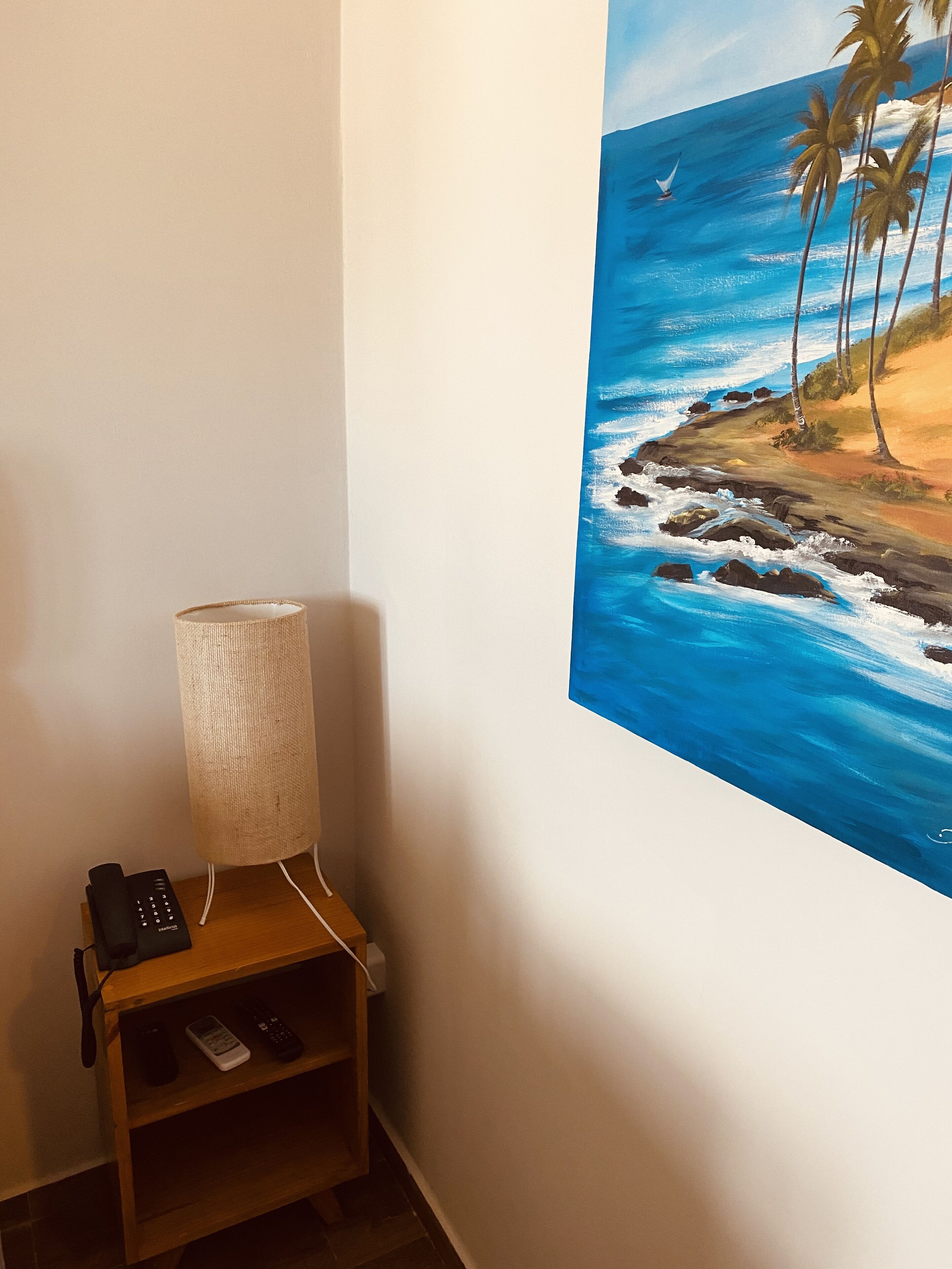 Una habitación con un cuadro colgado en la pared que muestra una escena de playa, una lámpara y un estante con controles remotos.