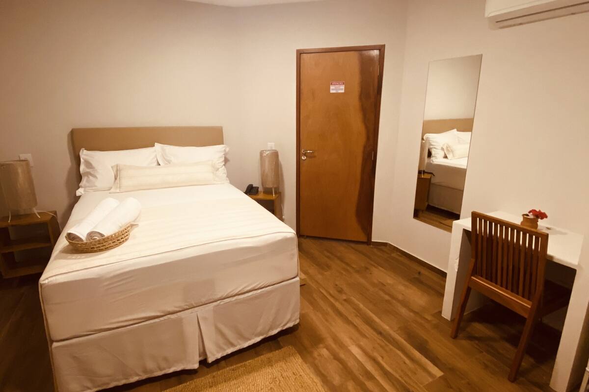 A hotel room with a bed, bedside tables, a desk, a chair, a wooden floor, a door, and a mirror.