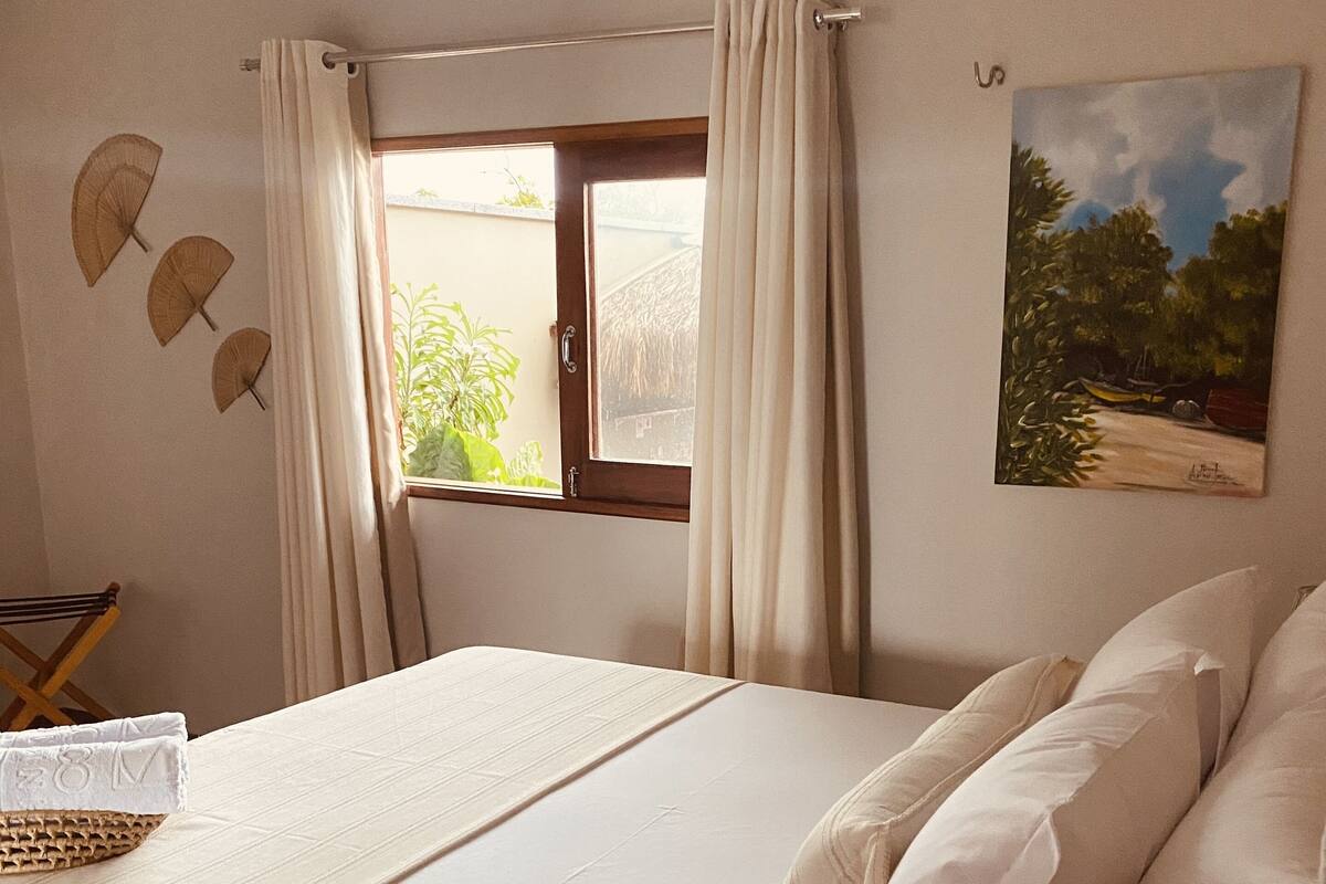A neatly made bed with white linens and pillows, a wooden side table with a glass container, a window with curtains, and a painting on the wall.