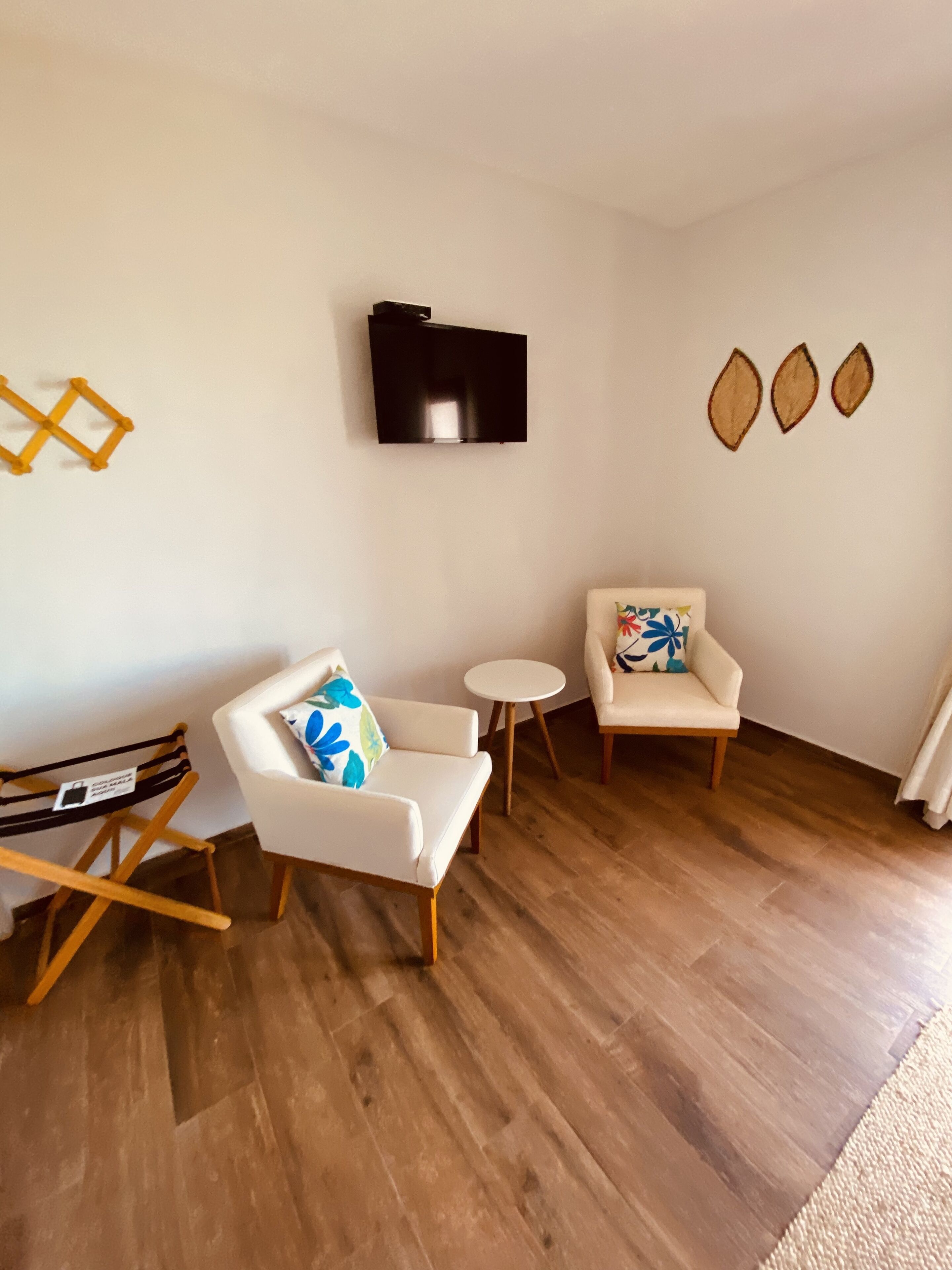 A living room with a wooden floor, a sofa with a blue and white pillow, a chair with a floral pillow, a small round table, and a flat-screen TV mounted on the wall.