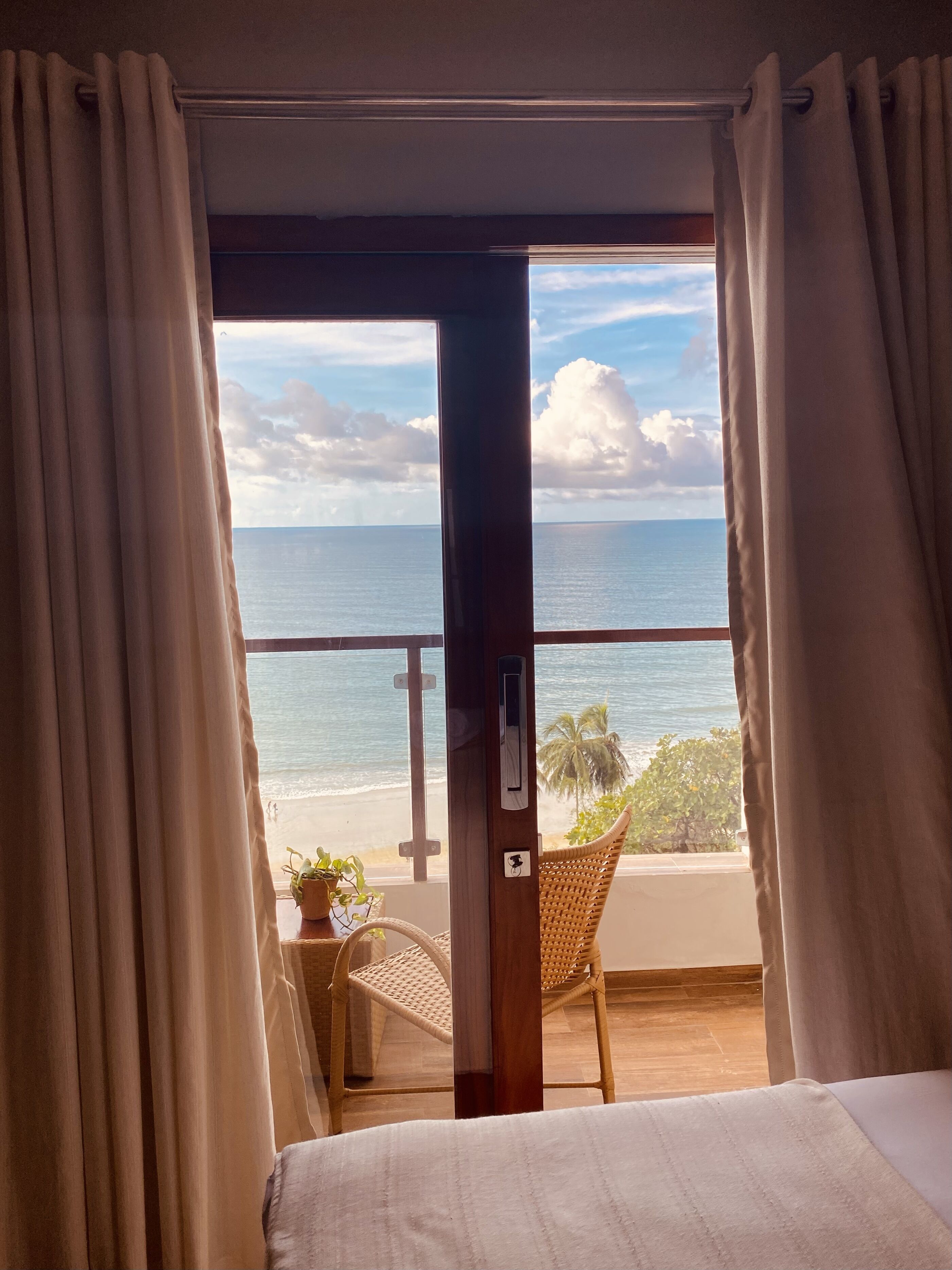 A room with a bed, a chair, and a view of the ocean through a window with curtains.