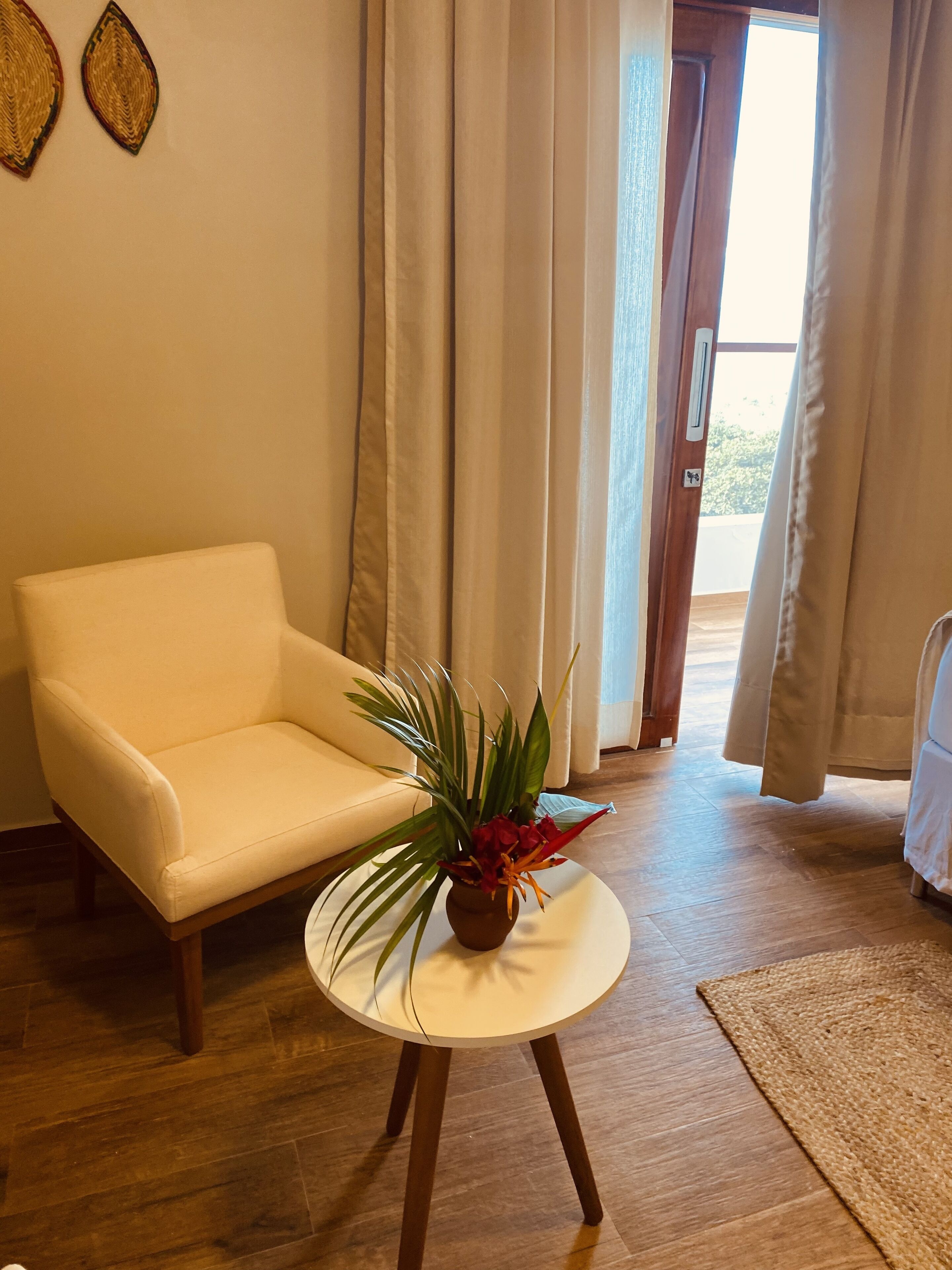 Una habitación con un sofá blanco, una mesita redonda con una planta, suelo de madera y vistas a través de una puerta con cortinas.