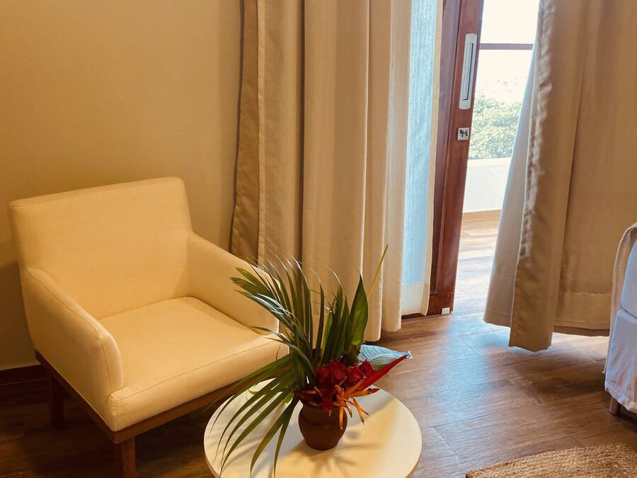 Una habitación con un sofá blanco, una mesita redonda con una planta, suelo de madera y vistas a través de una puerta con cortinas.
