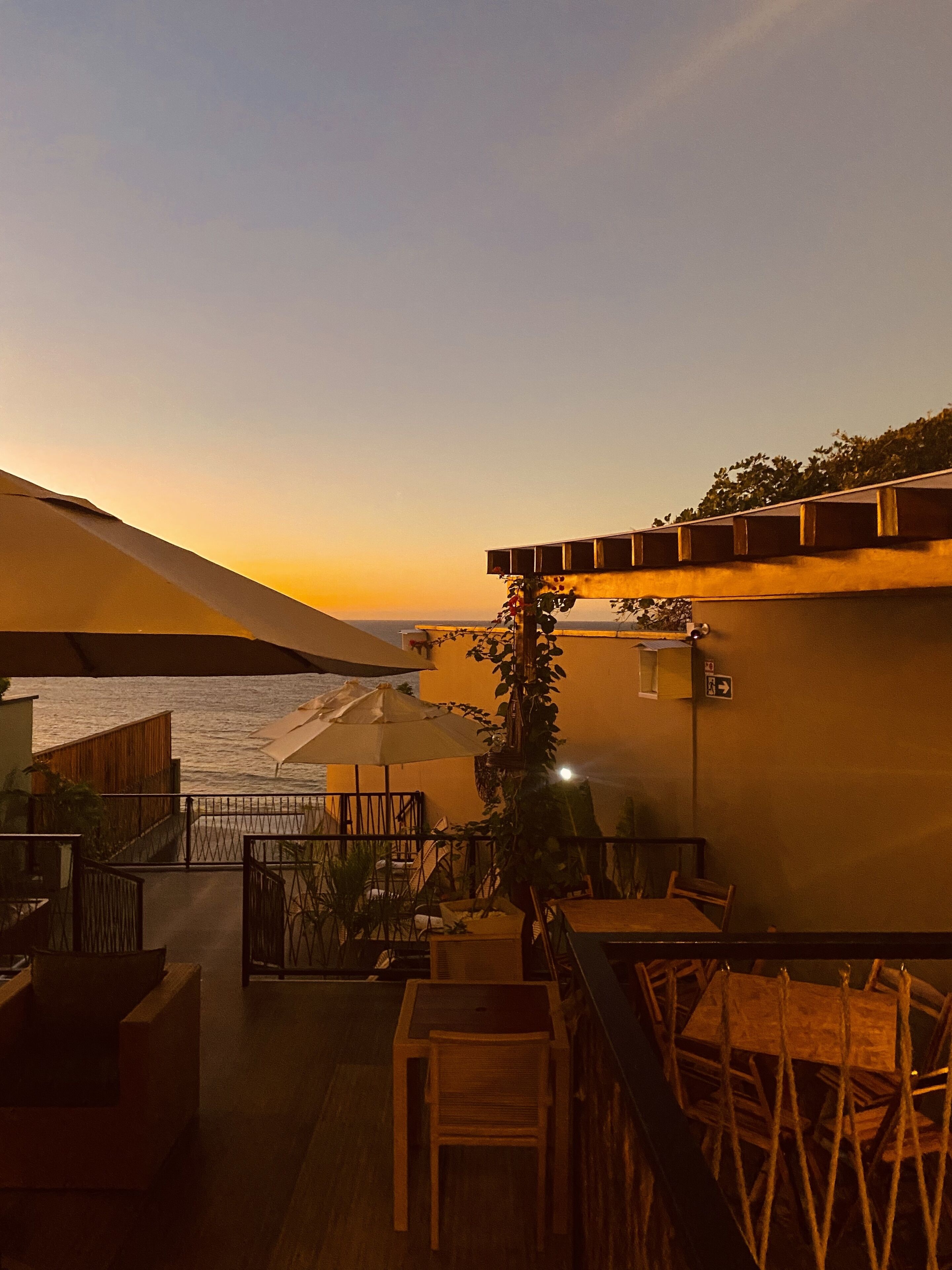A terrace with a view of the sea at sunset, featuring outdoor seating and a railing.