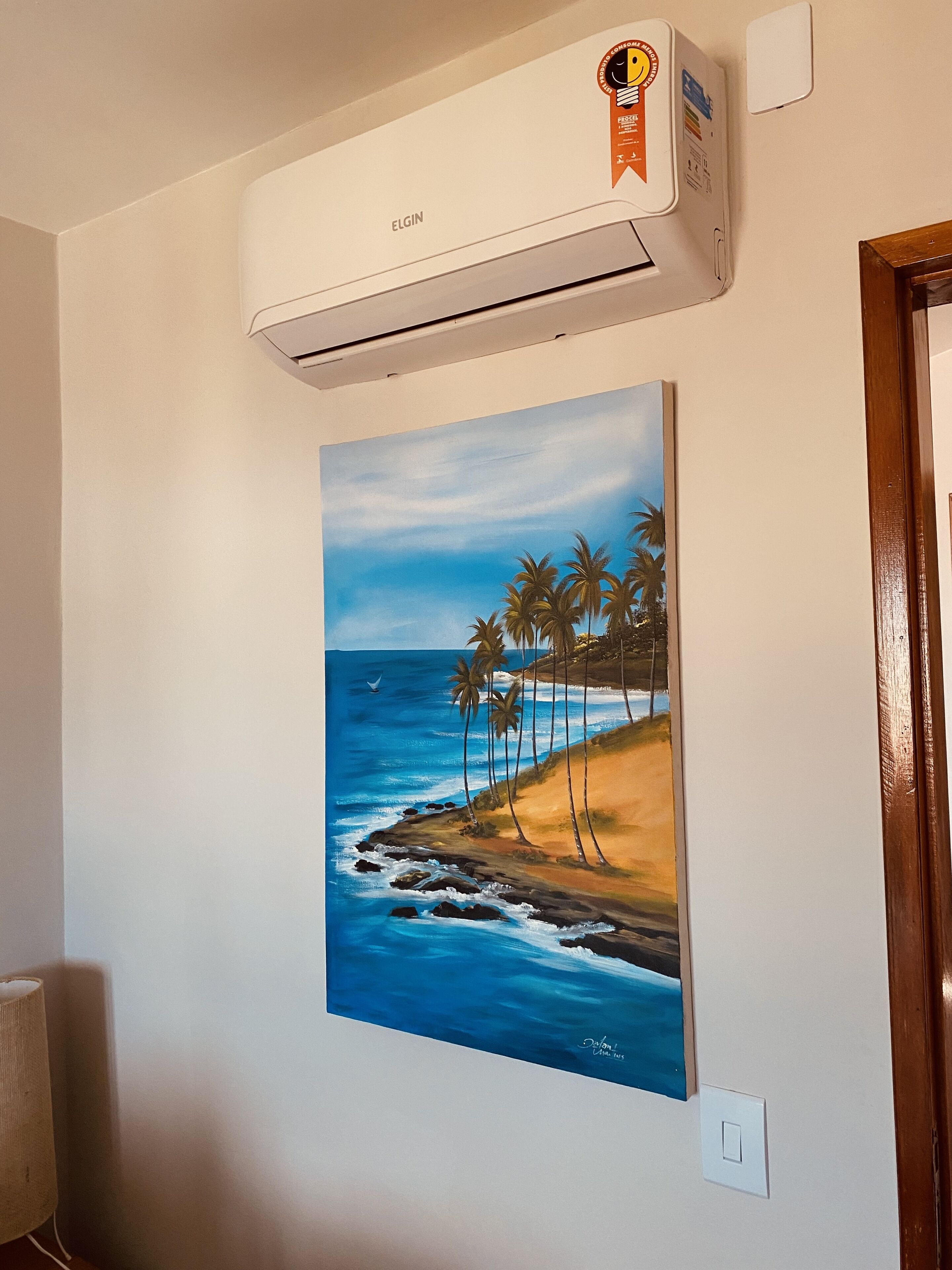 Een luchtverkoeler aan de muur, een sticker, een schilderij van een strand en een lichtschakelaar.