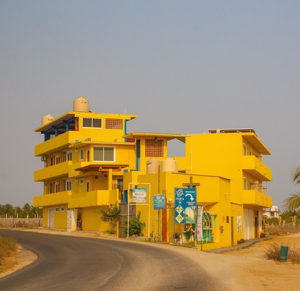 Un edificio amarillo de varios pisos con balcones, un tanque de agua en el techo y varias señales, incluyendo una de playa.