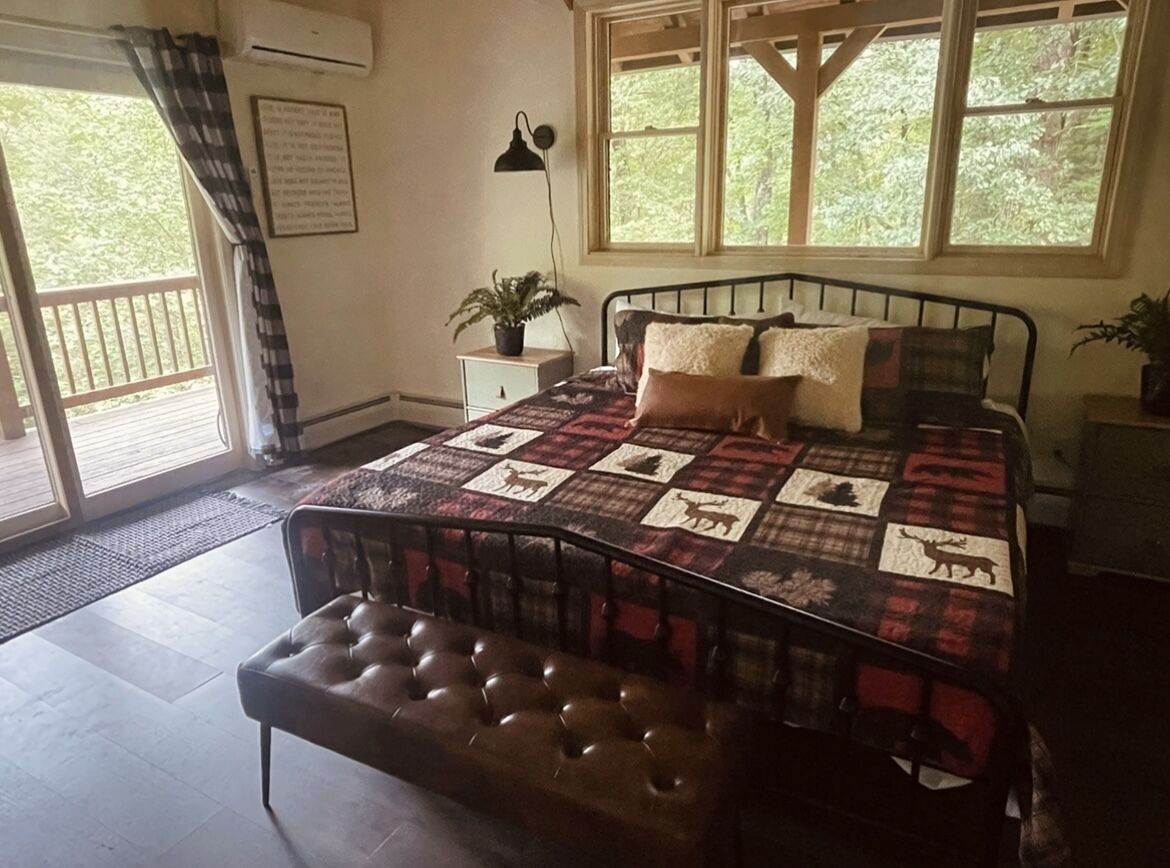 A bedroom with a bed, a bench, a nightstand, a potted plant, and a window with a view of trees.