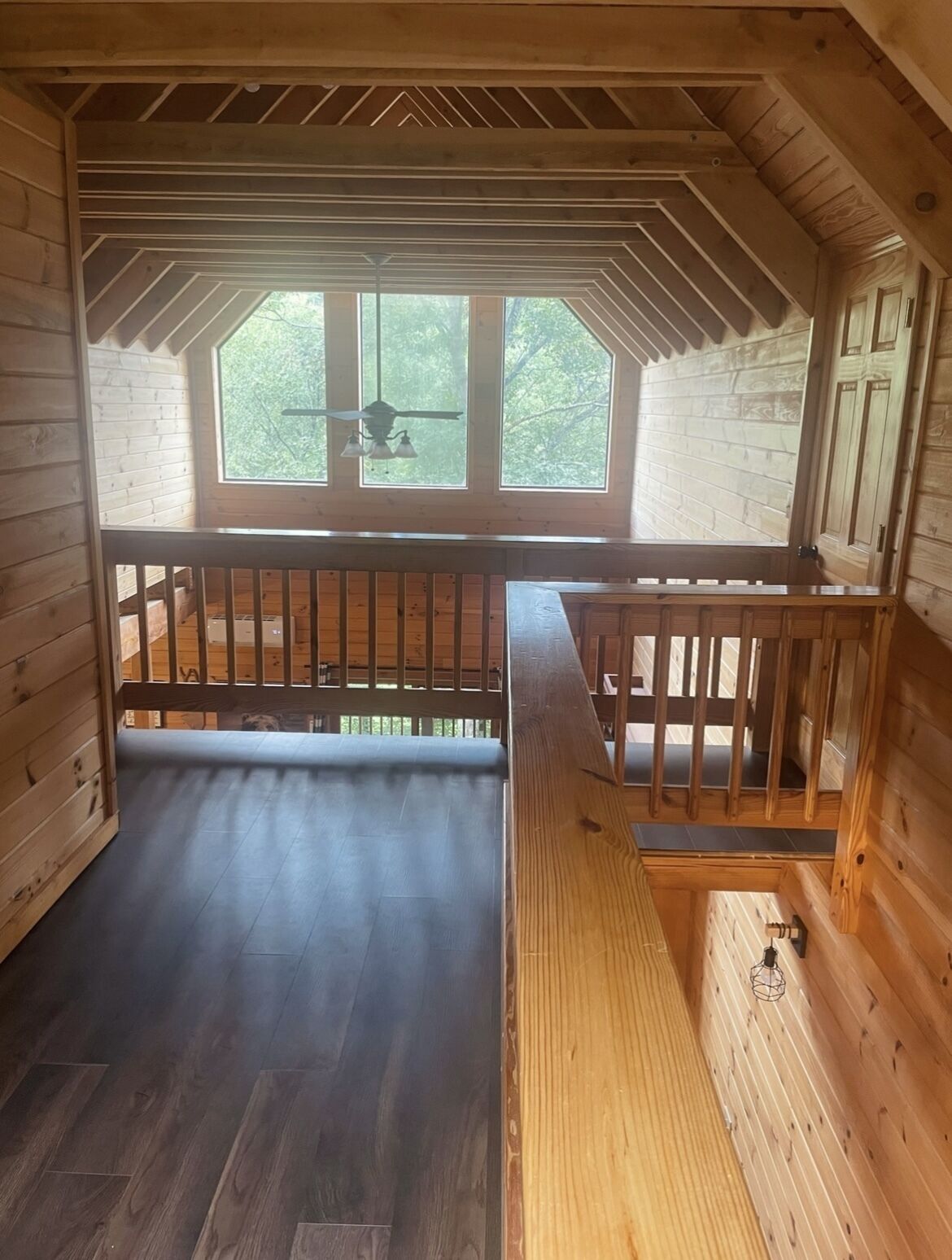 A wooden cabin with a loft bedroom, a balcony, and a large window providing a view of the outdoors.