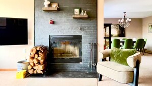 Una sala de estar con chimenea, un televisor montado en la pared y una zona de comedor con sillas verdes.