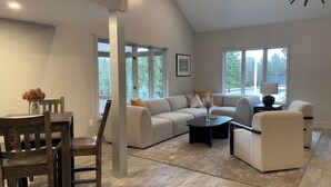 Una sala de estar moderna con un sofá seccional, una mesa de centro y un comedor con mesa y sillas de madera. Amplias ventanas ofrecen luz natural y vistas al exterior.
