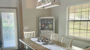 Un espace repas avec une table en bois, des chaises blanches, un luminaire suspendu et un panneau d’affichage en forme d’ardoise.