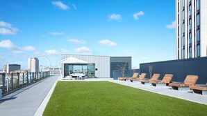 A rooftop terrace with a grassy area, lounge chairs, and a modern building with large windows. - Trendy South Bank 1BR w/ Pool, Gym & Concierge, nr Tube, by Blueground (London)