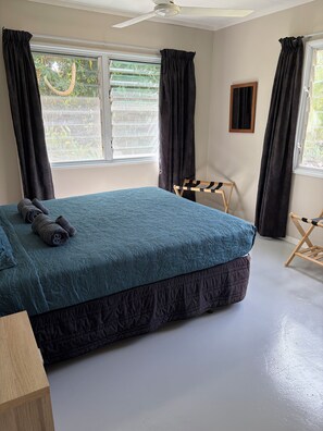 Ein Schlafzimmer mit einem Bett, einem Deckenventilator, einem Fenster mit Vorhängen und einem kleinen Holzstuhl.