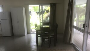 A room with a dining table and chairs, a white refrigerator, and a sliding glass door leading to a balcony with plants.