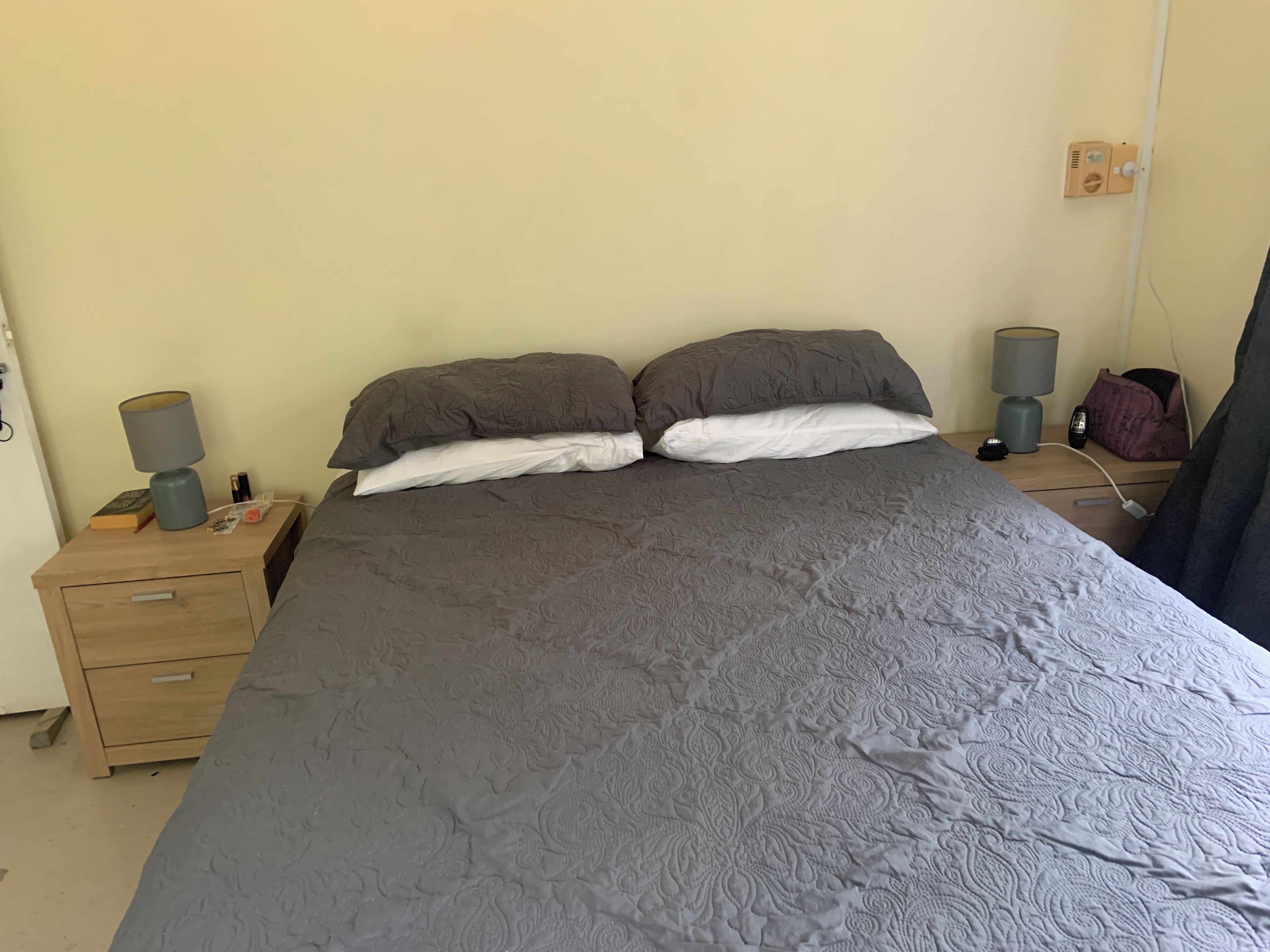 Un lit avec une couette grise, deux oreillers, une table de chevet avec une lampe et une prise de courant.