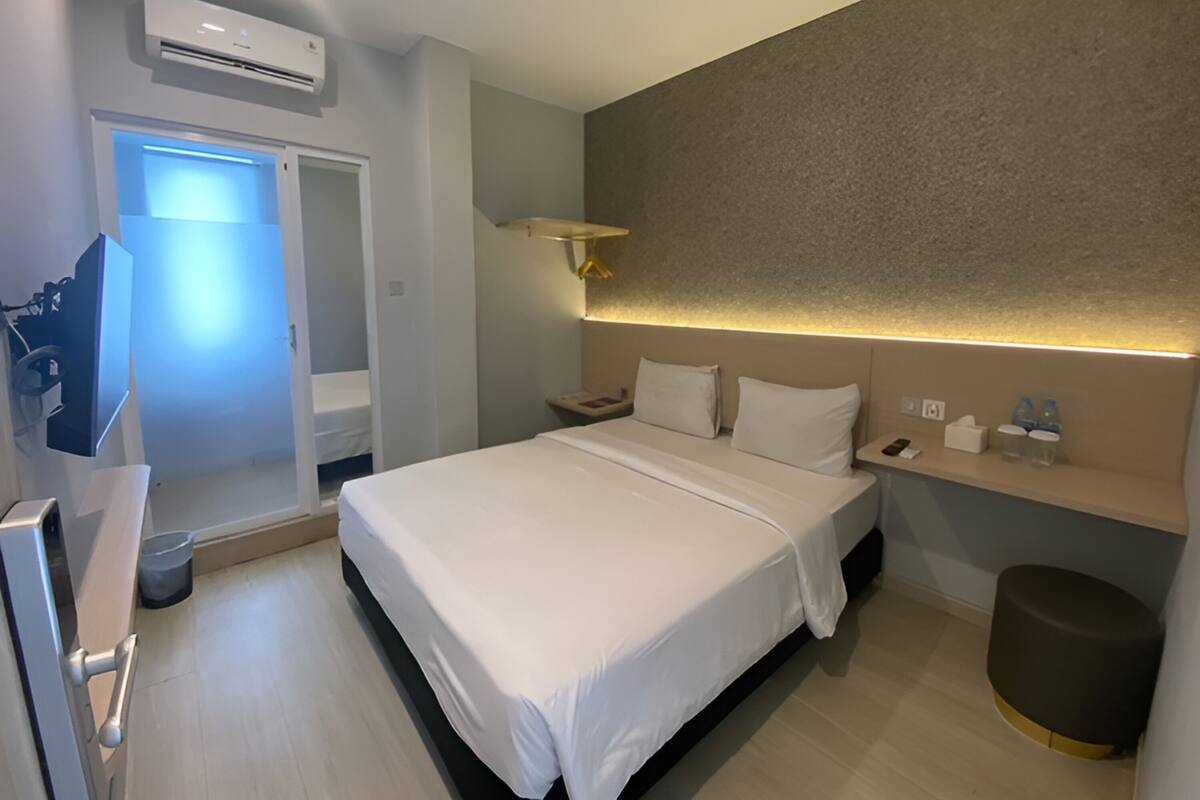 A modern hotel room with a neatly made bed, a bedside table with a lamp, a wall-mounted shelf with toiletries, and a door leading to a bathroom.