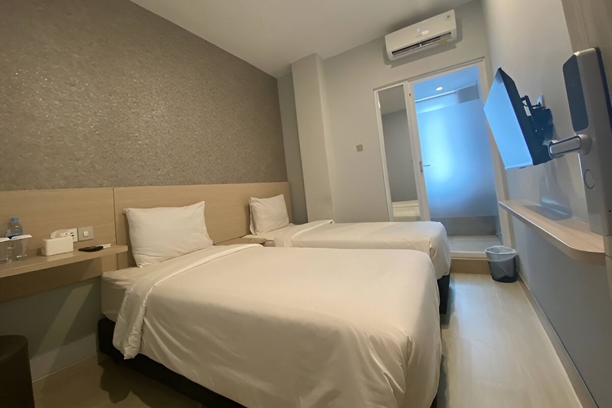 A hotel room with two beds, a bedside table, a mirror, an air conditioning unit, and a door leading to another room.