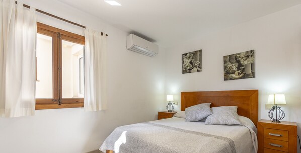 A bedroom with a bed, two bedside tables with lamps, a window with curtains, and two wall-mounted paintings.
