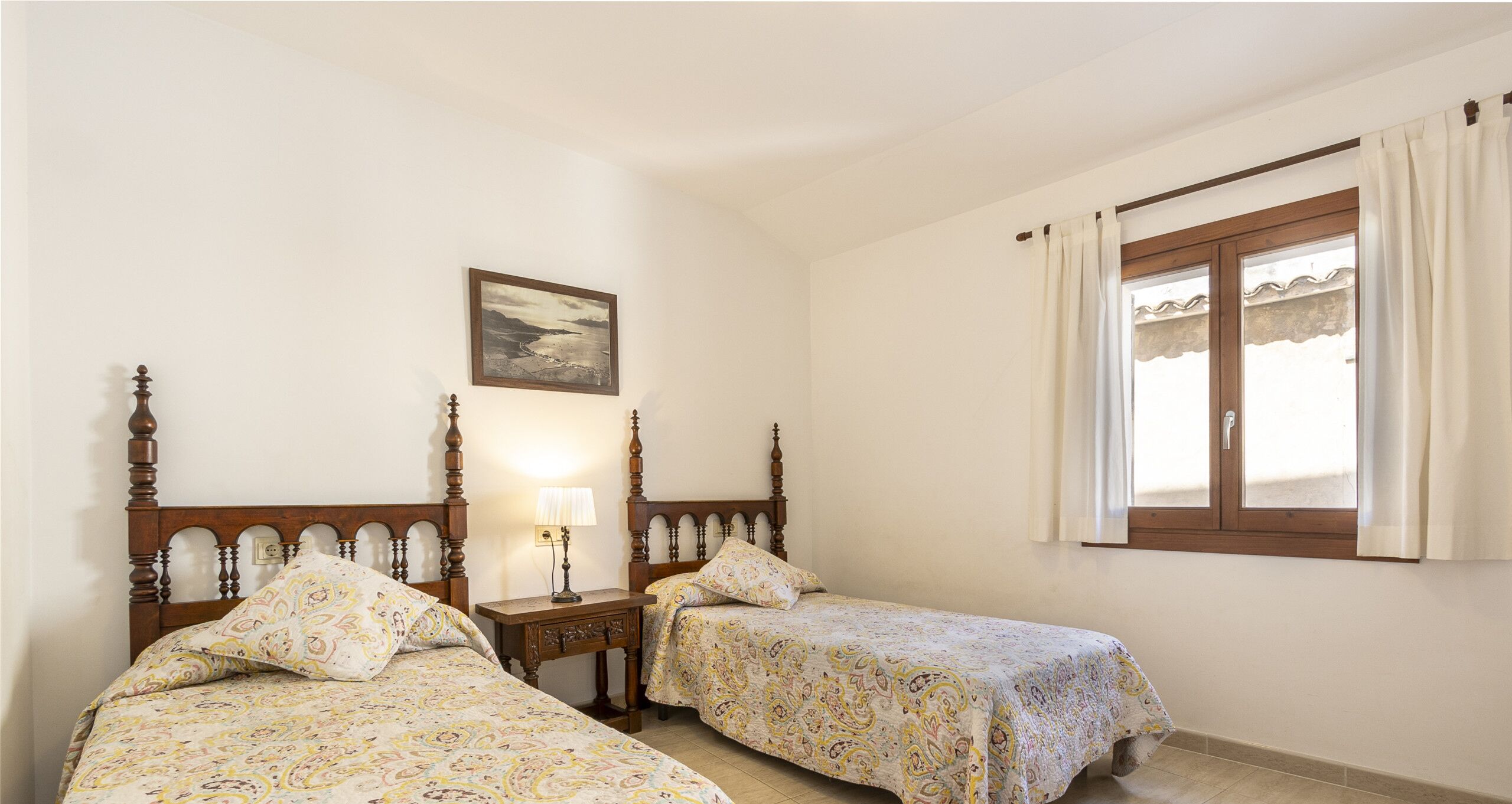 A bedroom with two wooden beds, a nightstand with a lamp, a window with curtains, and a framed picture on the wall.