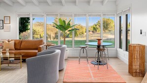 Ein modernes Wohnzimmer mit einer Ledersofa, grauen Sesseln, einem Couchtisch und einem Essbereich mit einem runden Tisch und Hockern. Große Fenster bieten Blick auf Bäume und eine Wiese.