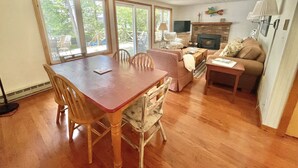 A spacious living room with a wooden dining table, comfortable seating, a fireplace, and large windows offering a view of the outdoors. - Waterfront, peaceful, cozy, cottage w/dock amazing sunsets, kayaks,3 bed/2 bath. (Lake Leelanau)