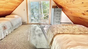 A cozy attic bedroom with two beds, a dresser, and a sliding glass door leading to a balcony. - Waterfront, peaceful, cozy, cottage w/dock amazing sunsets, kayaks,3 bed/2 bath. (Lake Leelanau)