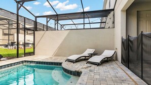 A swimming pool with two lounge chairs, a covered patio area, and a view of a building in the background.