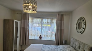 A bedroom with a bed, a wardrobe, a window with curtains, a hanging light fixture, and a wall mirror. - Town Centre Studio - Sleeps 2 - Parking (Reading)