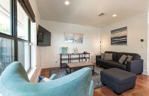 Una sala moderna con sofá, reposapiés y una mesa de centro. Hay un televisor montado en la pared, un cuadro y una ventana que deja entrar luz natural.