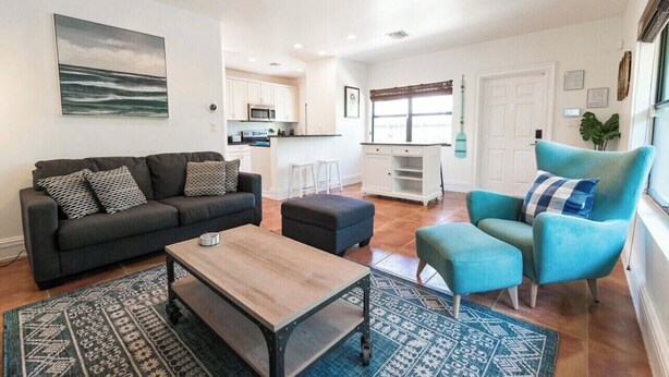Een moderne woonkamer met een bank, een poef, en een fauteuil, een salontafel, en een keuken op de achtergrond.