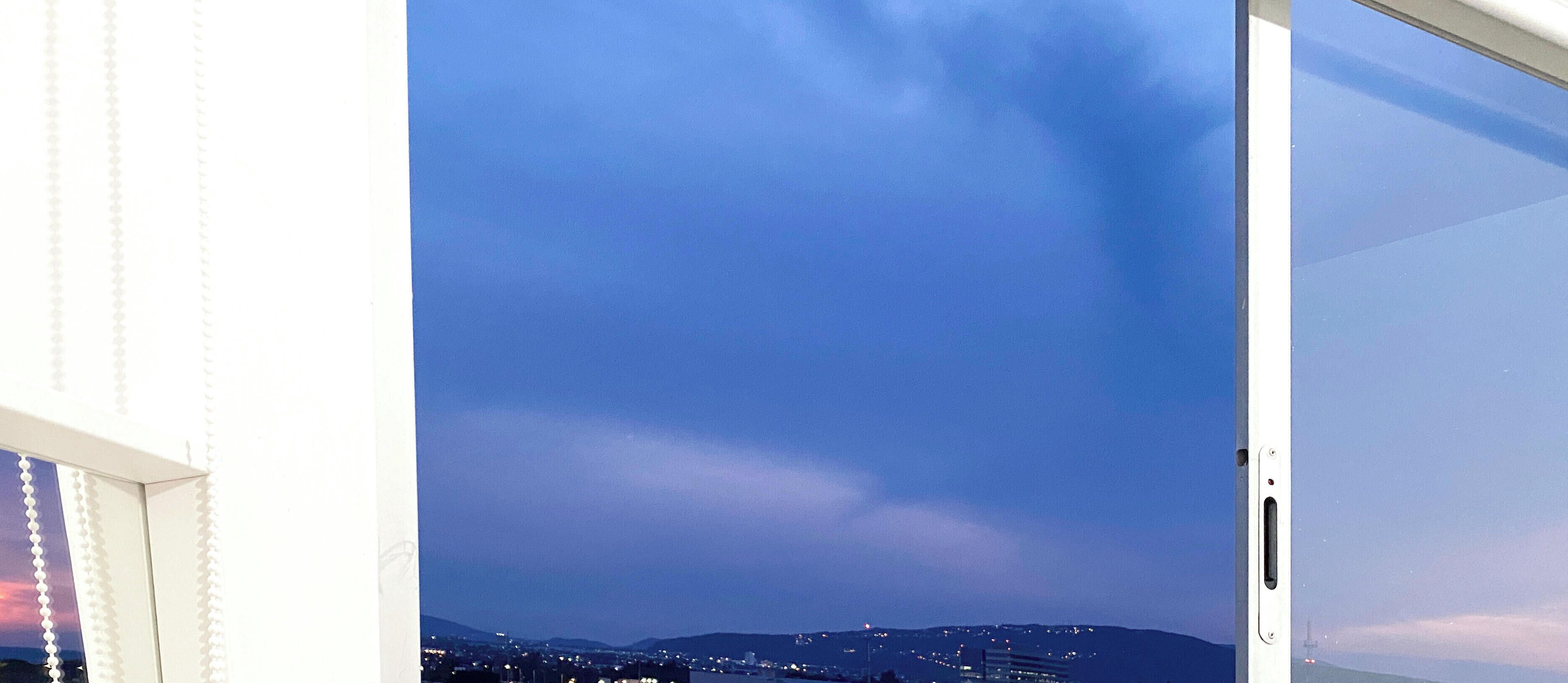 Blick durch ein offenes Fenster auf eine Stadtansicht im Abenddämmerung mit Gebäuden und einer entfernten Hügelkette.