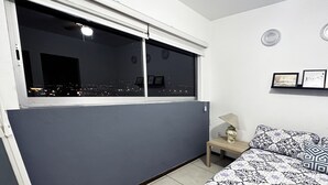 Une pièce avec une grande fenêtre, un lit recouvert d’une literie à motifs, une petite table de chevet avec une lampe, et deux tableaux encadrés accrochés au mur.