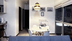 Un salon moderne avec un canapé bleu, une table à manger et des chaises, ainsi qu’un lustre. Des tableaux encadrés ornent le mur et de grandes fenêtres offrent une vue sur la ville.