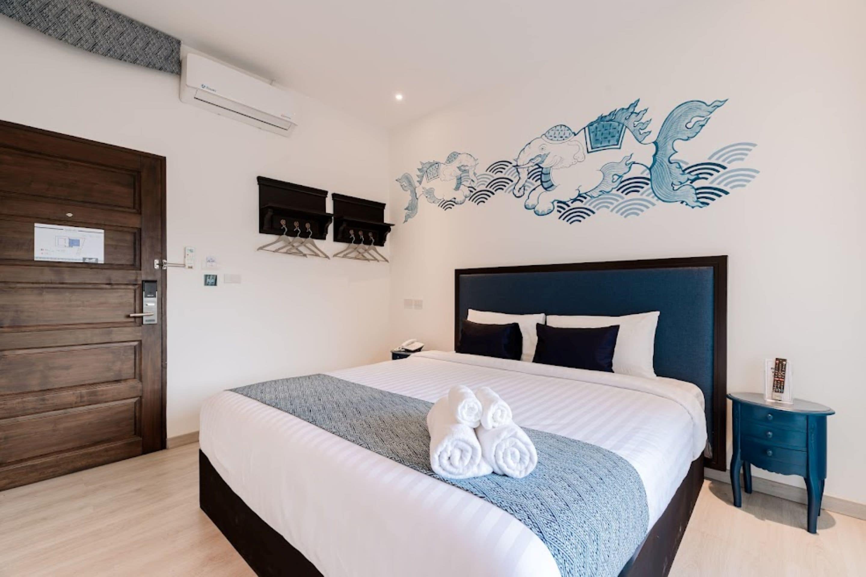 Una cama bien hecha con sábanas blancas y una manta azul, un aire acondicionado de pared, una puerta de madera, una mesita de noche pequeña con una lámpara y un mural decorativo en la pared.