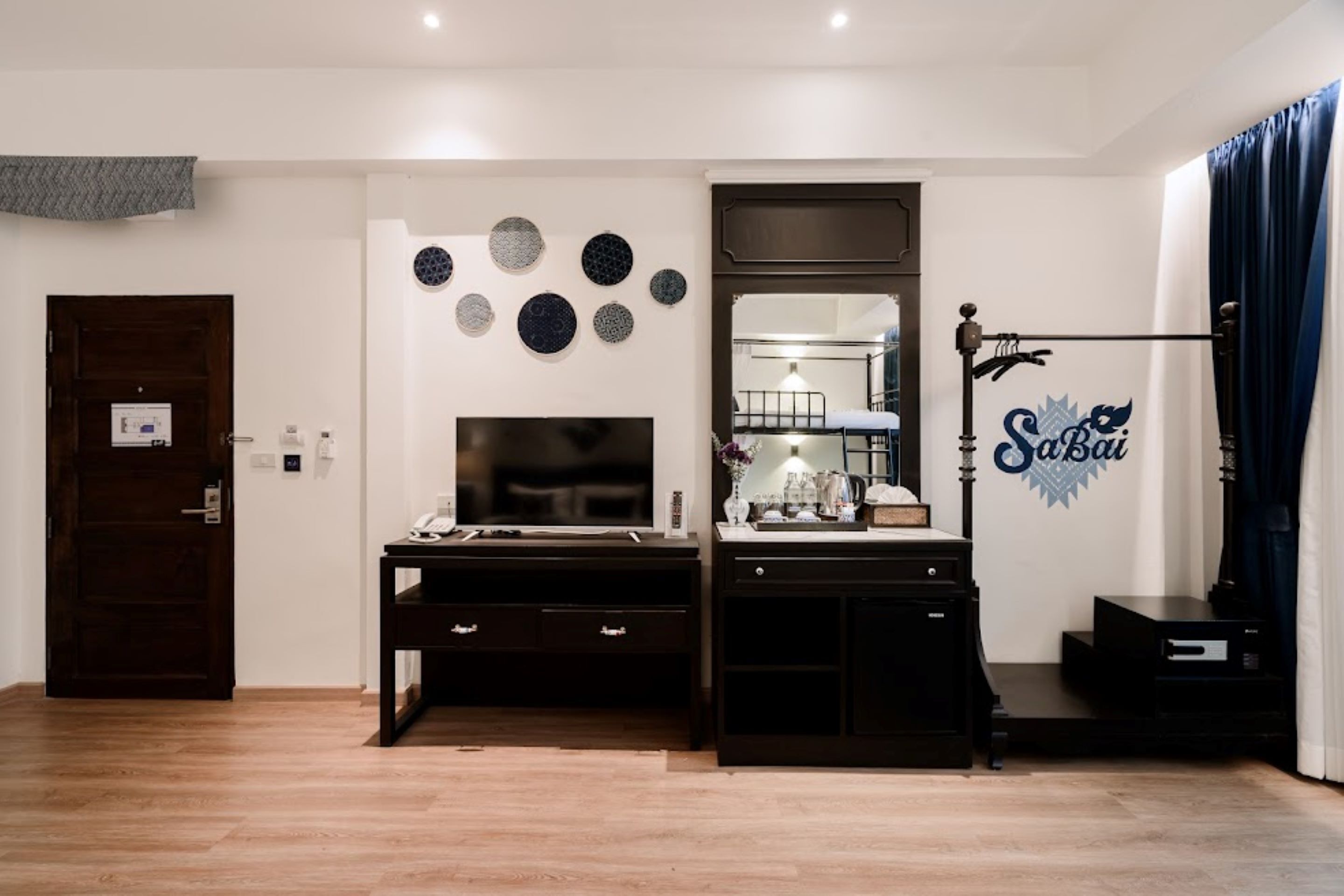 A modern room with a wooden floor, a dark desk with drawers, a large flat-screen TV, a mirror, and a decorative wall with circular patterns.